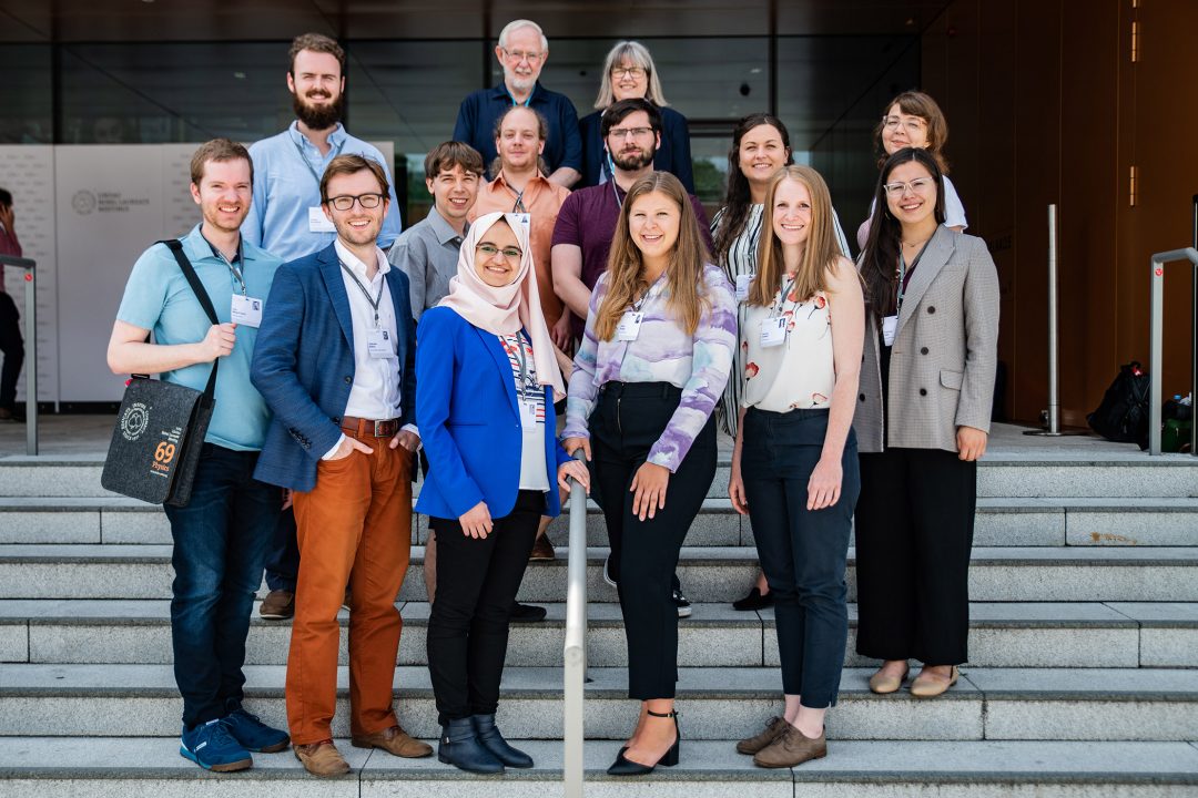12 young scientists standing on stairs smiling into the camera. Canadian Nobel Laureates Arthur McDonald and Donna Strickland are standing behind them.